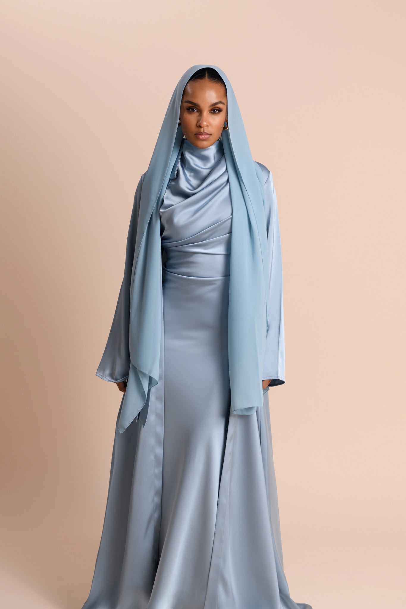 Woman wearing a long, light blue hijab and matching dress against a beige background