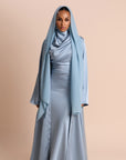 Woman wearing a long, light blue hijab and matching dress against a beige background