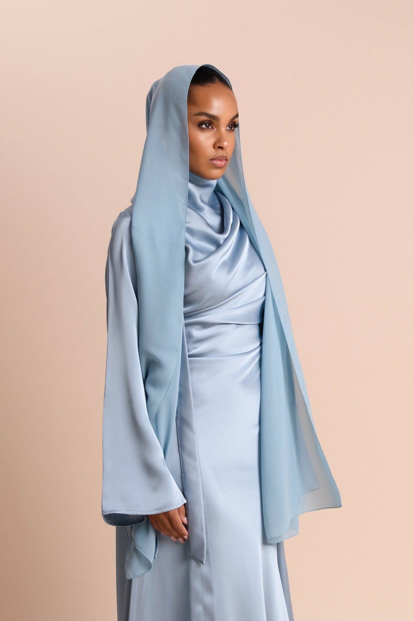 Woman wearing a light blue hijab and dress against a beige background